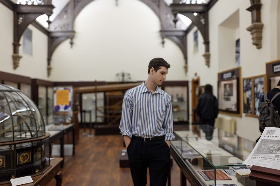 Alt Text: Landscape photo where John Kirkley walks through the Whipple Museum, surrounded by historical artefacts and gothic architecture. He looks away from the camera. He is wearing a blue striped shirt and has brown hair.