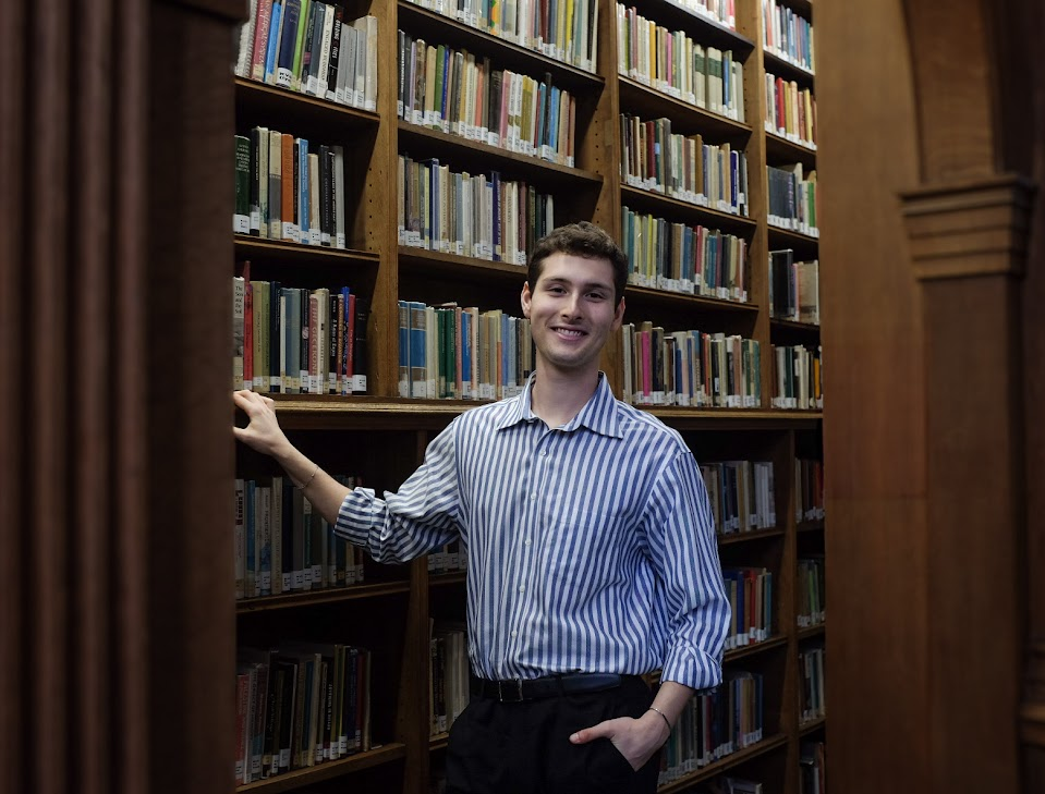 Alt Text: Landscape photo where John Kirkley smiles directly at the camera. He is lightly holding a bookshelf, and has the other hand in his pocket. He is surrounded by an archway and bookshelves in the Haddon Library. He is wearing a blue striped shirt a