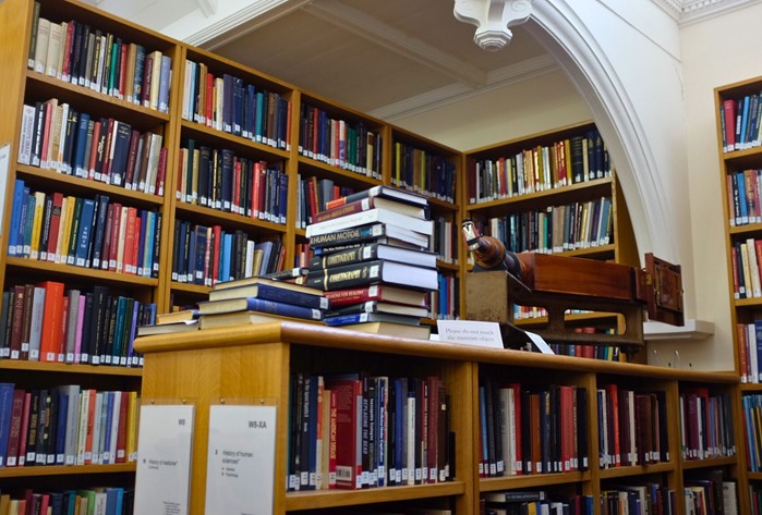 Photo of tall library shelves filled with books, with further books stacked in a pile in front of them. 