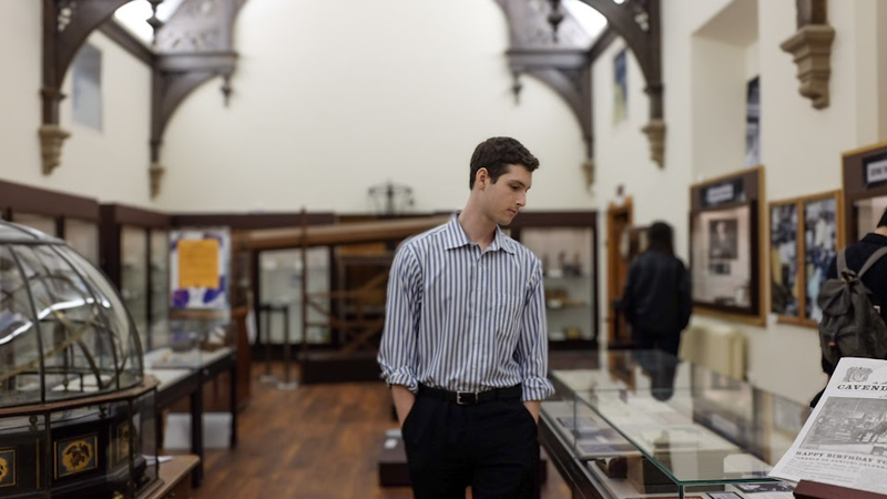 Alt Text: Landscape photo where John Kirkley walks through the Whipple Museum, surrounded by historical artefacts and gothic architecture. He looks away from the camera. He is wearing a blue striped shirt and has brown hair.
