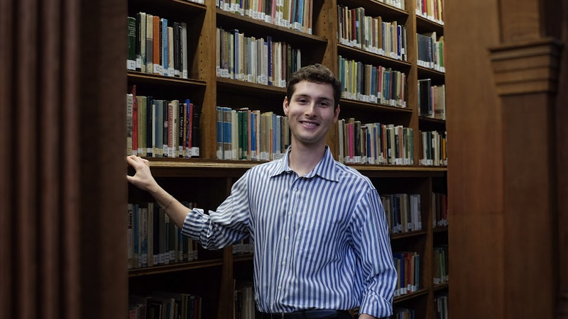Alt Text: Landscape photo where John Kirkley smiles directly at the camera. He is lightly holding a bookshelf, and has the other hand in his pocket. He is surrounded by an archway and bookshelves in the Haddon Library. He is wearing a blue striped shirt a