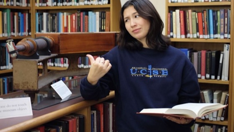 Photo of a student gesturing at a display in the Whipple Library, while holding a book open in the other hand.