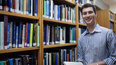 Alt Text: Portrait photo where John Kirkley smiles directly at the camera. He is holding a book, and is surrounded by bookshelves in the Whipple Library. He is wearing a blue striped shirt and has brown hair.