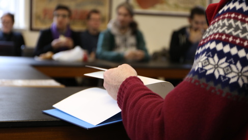 Soft focus view of a seminar in progress, with a person in a patterned jumper flipping pages of a notebook in the foreground.