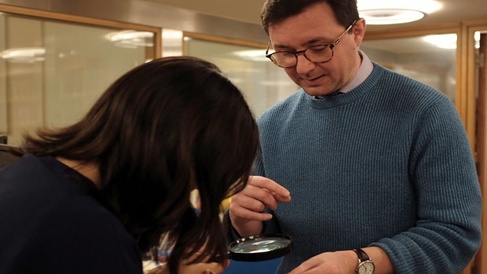 Photo of a student examining a tiny book, held out by the Librarian Jack Nixon, under a microscope,. 
