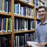 Alt Text: Portrait photo where John Kirkley smiles directly at the camera. He is holding a book, and is surrounded by bookshelves in the Whipple Library. He is wearing a blue striped shirt and has brown hair.