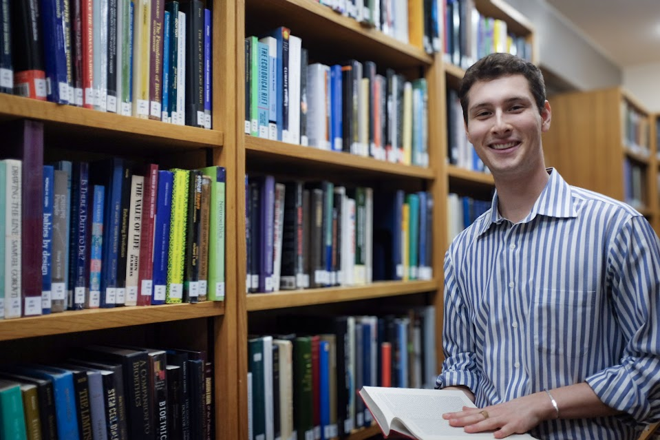 Photo taken by Farradeh Martin (c) Alt Text: Portrait photo where John Kirkley smiles directly at the camera. He is holding a book, and is surrounded by bookshelves in the Whipple Library. He is wearing a blue striped shirt and has brown hair.