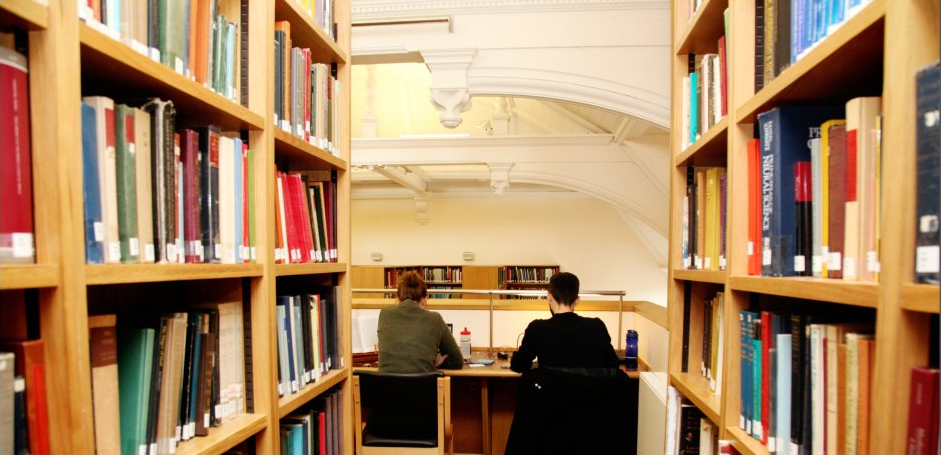 Two people working at a library desk, shown from behind and flanked by bookshelves.