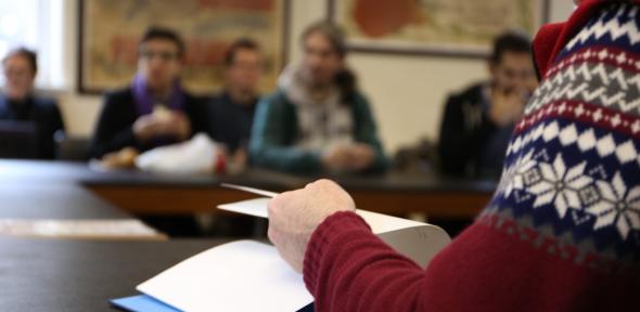 Soft focus view of a seminar in progress, with a person in a patterned jumper flipping pages of a notebook in the foreground.